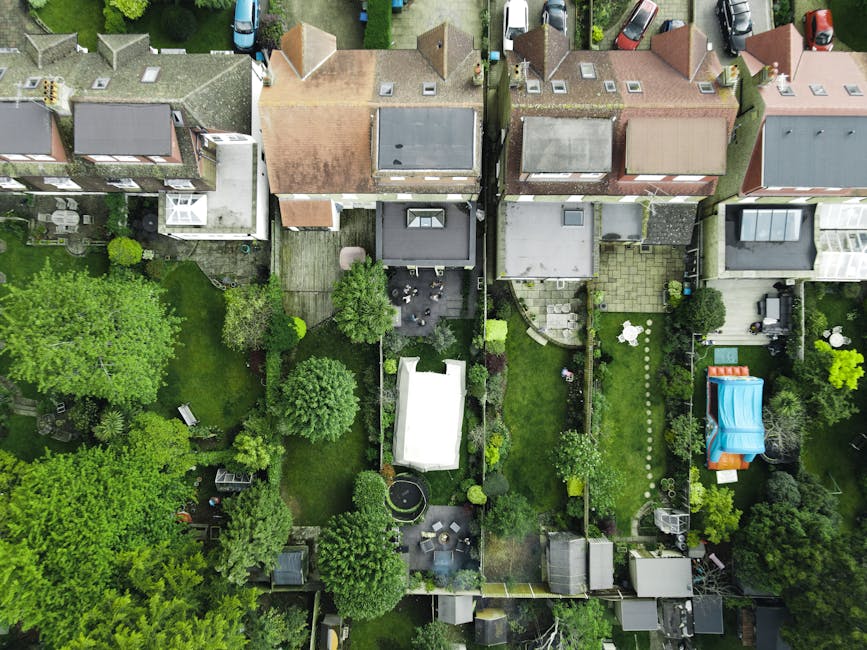 An aerial view of a row of suburban houses shows neatly maintained gardens with lush green lawns, mature trees, and various shrubs. The back gardens feature well-defined borders, paved patios, and gar